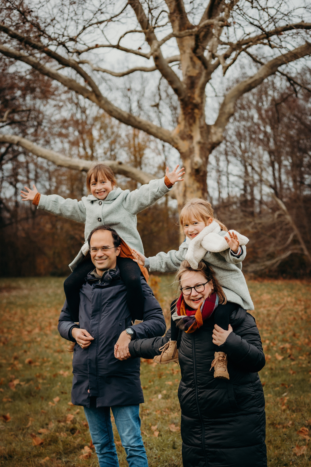 Zwei Mädchen sitzen auf den Schultern der Eltern und strecken die Arme nach oben
