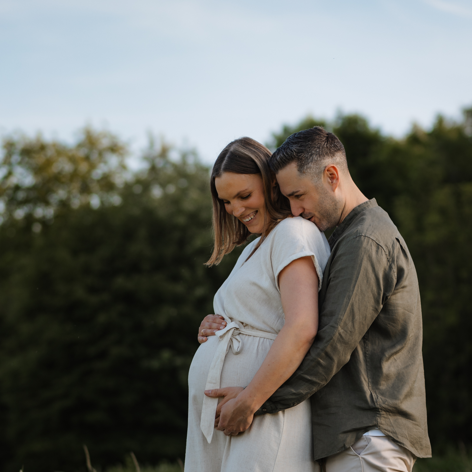Mann umarmt seine schwangere Frau liebevoll, während beide lächelnd auf den Babybauch schauen
